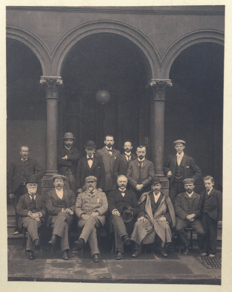 A sepia photograph of a group of 13 men and one boy, some seated, outside the Arlington Baths in 1896, dressed in suits and wearing caps or bowler hats