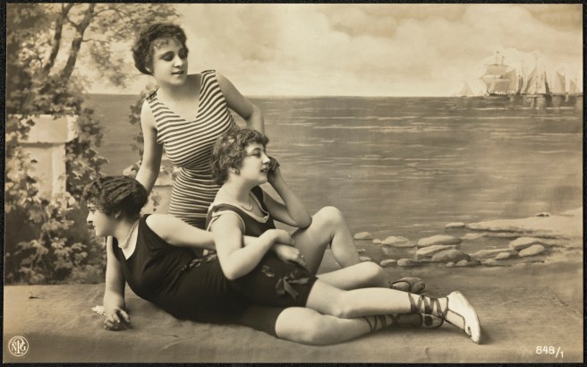 A sepia postcard with three young women in close-fitting swimsuits posing in a studio with a painted backdrop of the sea and boats. The swimsuits have no sleeves, V-necks and long shorts