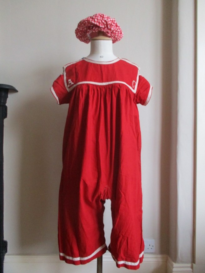 Photo of a replica swimming costume made of red cotton with short sleeves and bloomer-style legs displayed on a tailor’s dummy. It has white tape sewn around the edges of the chest yoke, the outside edges of the sleeves and around the bottom of the legs. A capital A and capital C are embroidered in the bottom corners of the chest piece. It has tape over the shoulders fastened with buttons at the front.