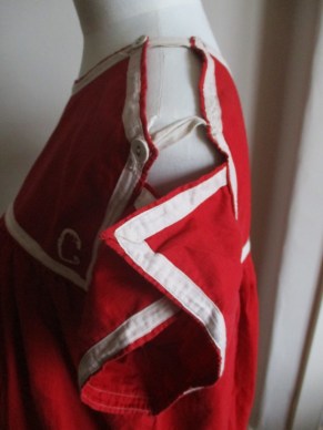 Photo of a replica swimming costume made of red cotton with short sleeves displayed on a tailor’s dummy, seen from the side and top. White tape from the back of the costume and buttoned to the front goes over the shoulders to hold the front and back together over. It has white tape sewn around the edges of the chest yoke and the outside edges of the sleeves. A capital C is embroidered in the visible bottom corner of the chest piece.