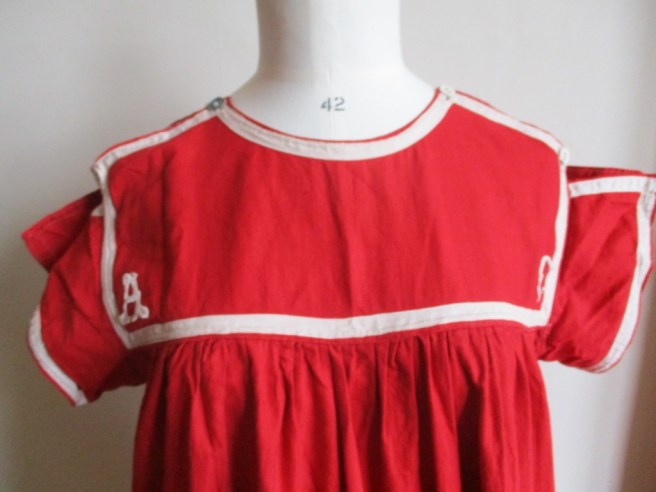 Photo of the chest of a replica swimming costume made of red cotton with white tape sewn around the edges of the chest yoke and the the outside edges of the sleeves. A capital A and capital C are embroidered in the bottom corners of the chest piece. It has tape over the shoulders fastened with buttons at the front