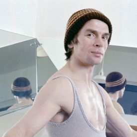 Colour photograph of Rudolf Nureyev in a sleeveless leotard and woolly hat, looking at the camera and his back reflected in two mirrors behind him