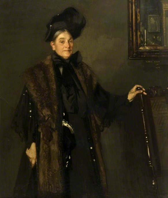 A portrait of a older woman in 19th century clothes: a long black dress, a black hat and a fox fur cape over her shoulders. She is standing with her hand on the back of an old chair with the corner of a painting behind her