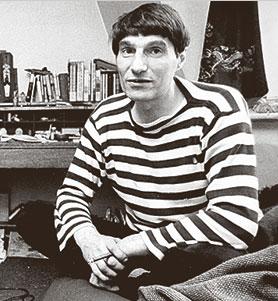 A black and white photo portrait of Alexander Trocchi in 1967, sitting crossed-legged and wearing a striped top. There is a desk and shelf of books behind him.