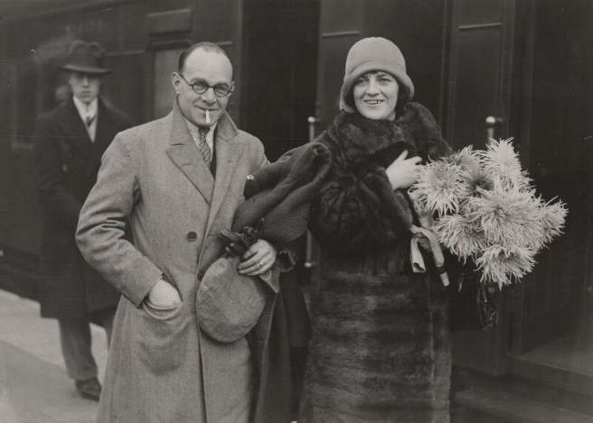 A black and white photo of a middle-aged man and woman on a railway platform with a train behind them.The man wears round glasses, a tweed overcoat and is smoking a cigarette and carrying leather gloves and a tweed cap. The woman is wearing a felt cloche hat, a long fur coat and is carrying a bag, gloves and a big bunch of chrysanthemums. Both are smiling.