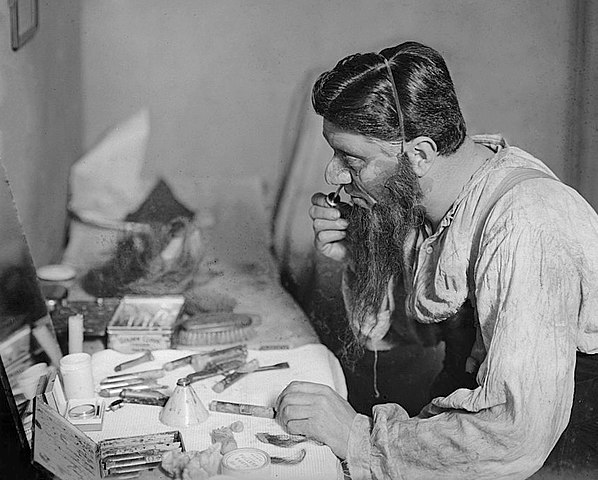 Bransby Williams seen in profile at a dressing table waeing a false beard and applying stage make-up.