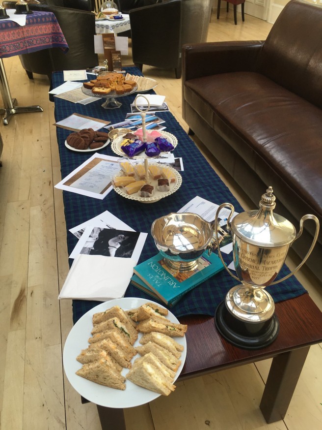 Table with plates of sandwiches and cakes, also photos, silver cups, books and copies of documents