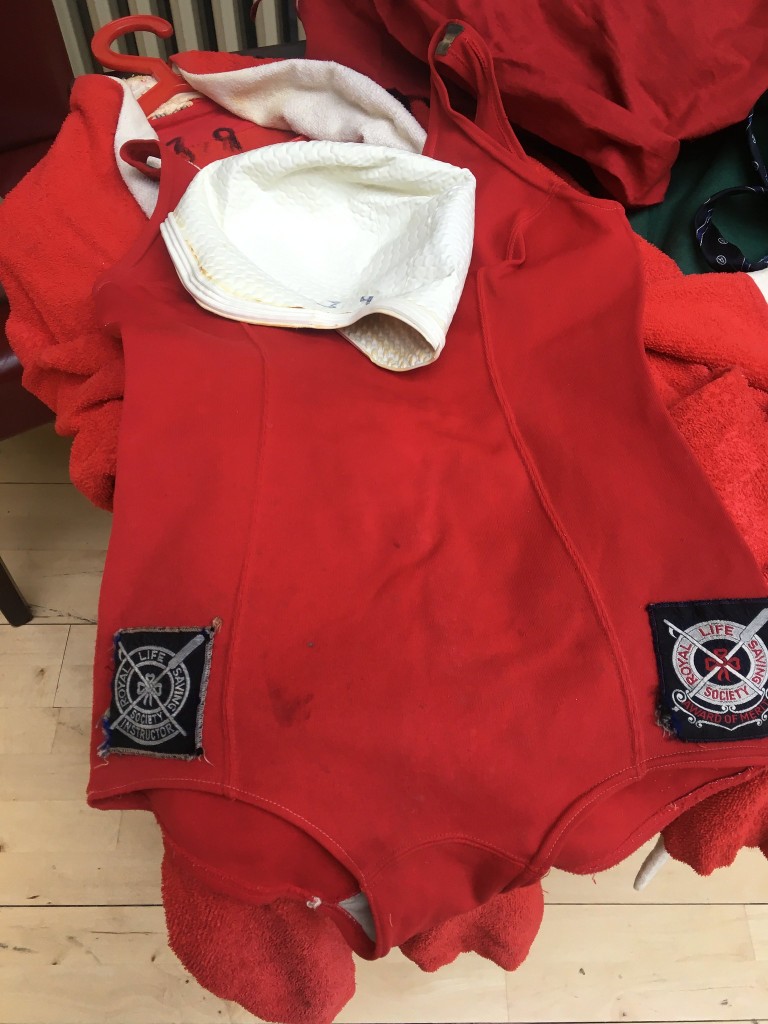Red swimming costume with lifesaving badges, a white swimming cap and a red towelling robe underneath