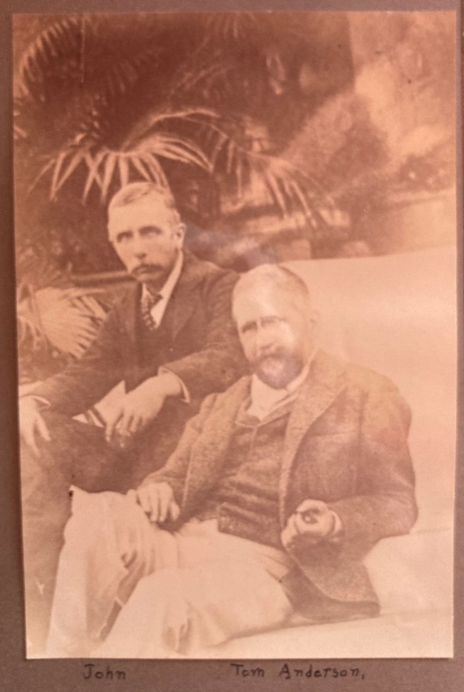 Sepia photo of two older men in Victorian clothes sitting down with palm leaves behind them. They are labelled 'John' and 'Tom Anderson'.