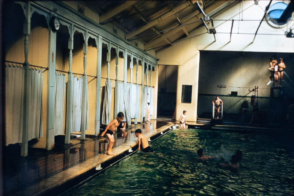 An old fashioned swimming pool with curtaned cubicles at one side and five boys either in the water or at the sides ready to dive in. At the end is one boy on a low springboard and other boys queuing up on a a higher diving platform. 